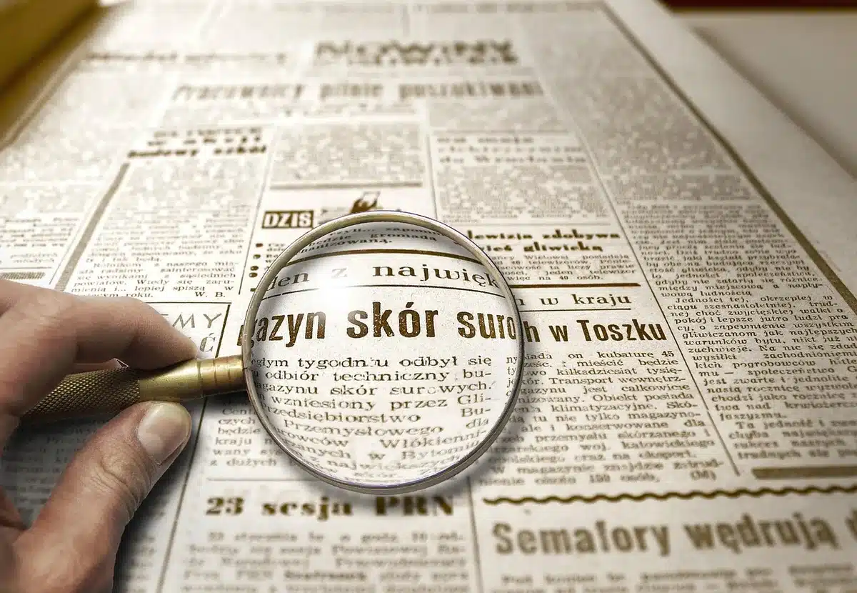 Hands holding a magnifying glass over an unbranded euro-style coin on a desk with a notepad and a pen nearby