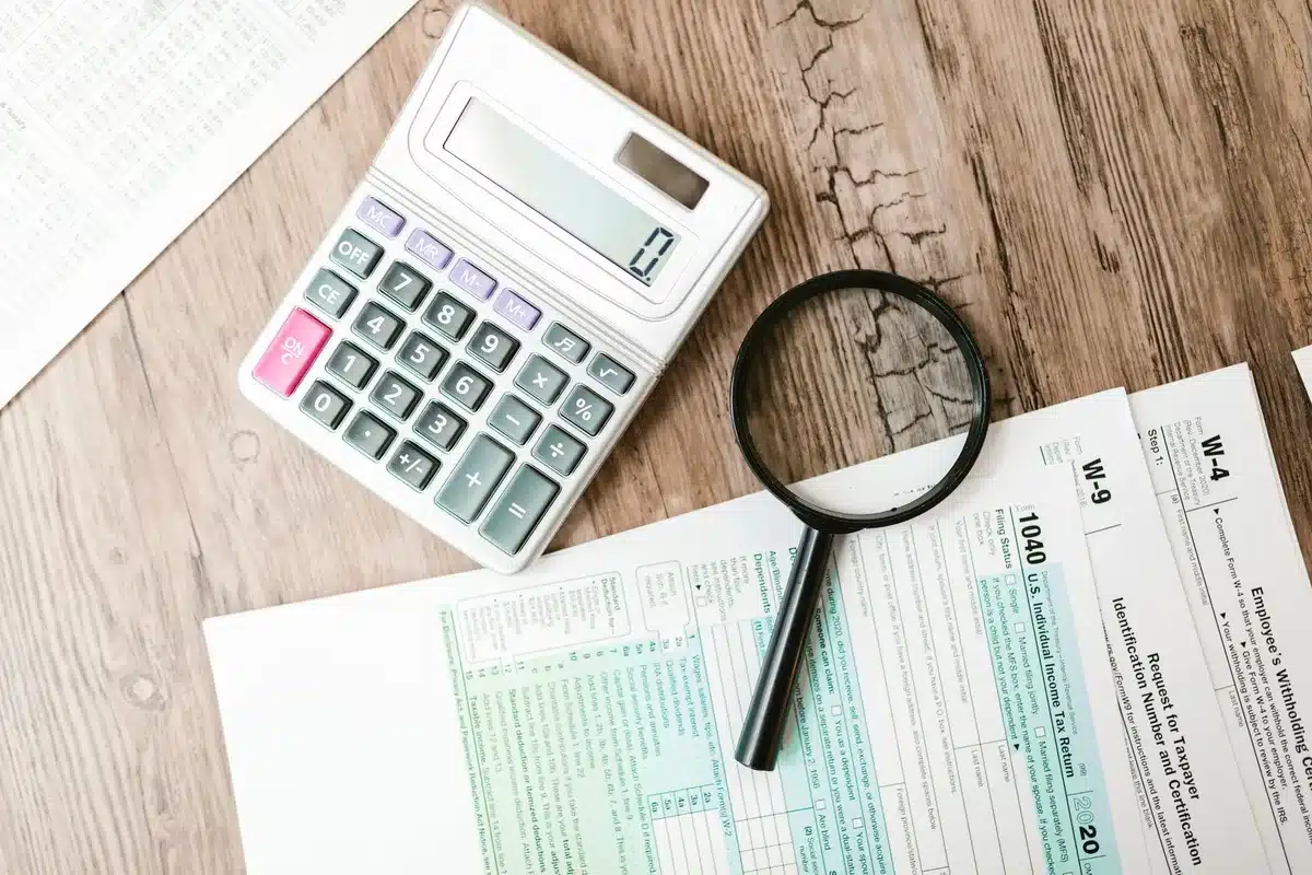 An open notebook on a desk with a pen, a calculator and a small stack of generic coins in the background