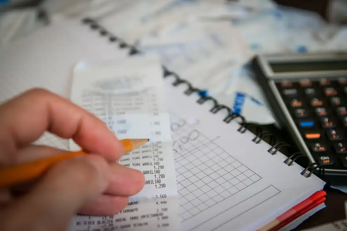 close-up of a hand holding a small jewellery receipt and a calculator on a light wooden counter, blurred gold items in background